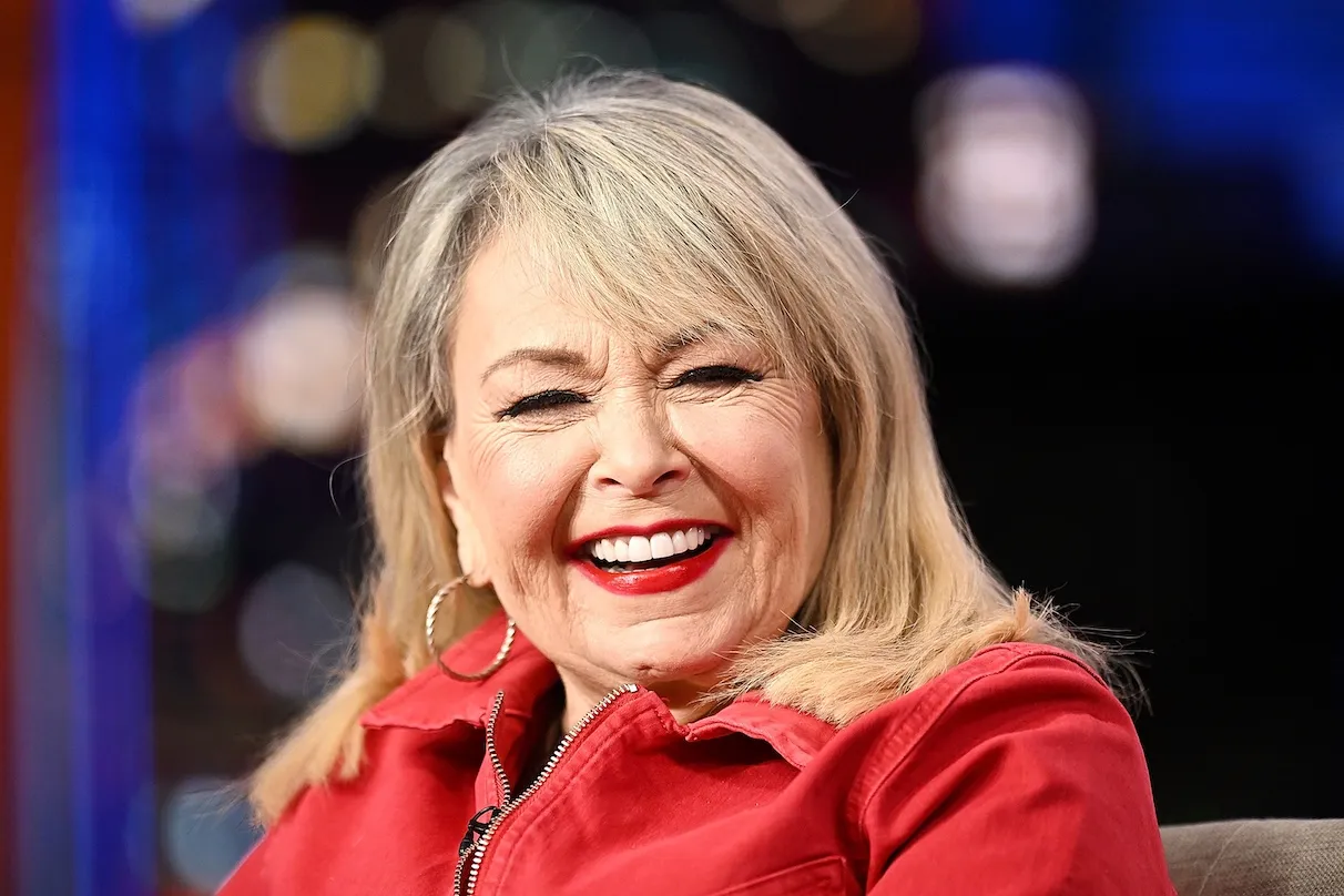 Roseanne Barr smiling and sitting. She's wearing a red jacket.