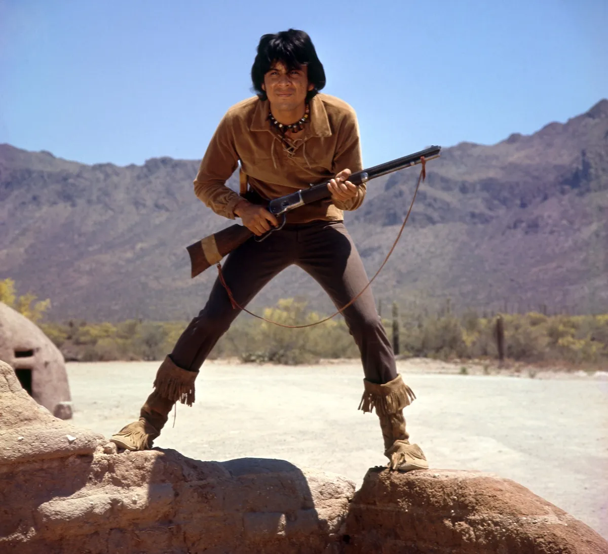 Rudy Ramos as Wind, holding a rifle and standing on a rock in 'The High Chaparral'