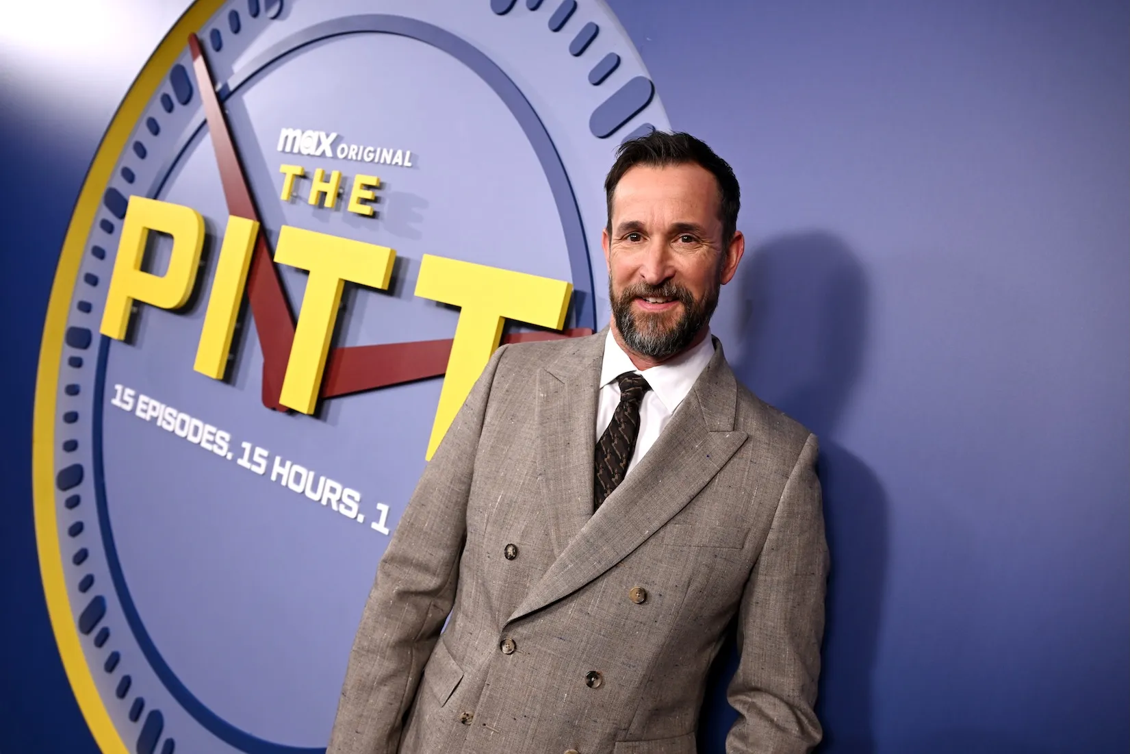 Noah Wyle standing in a suit in front of 'The Pitt' background