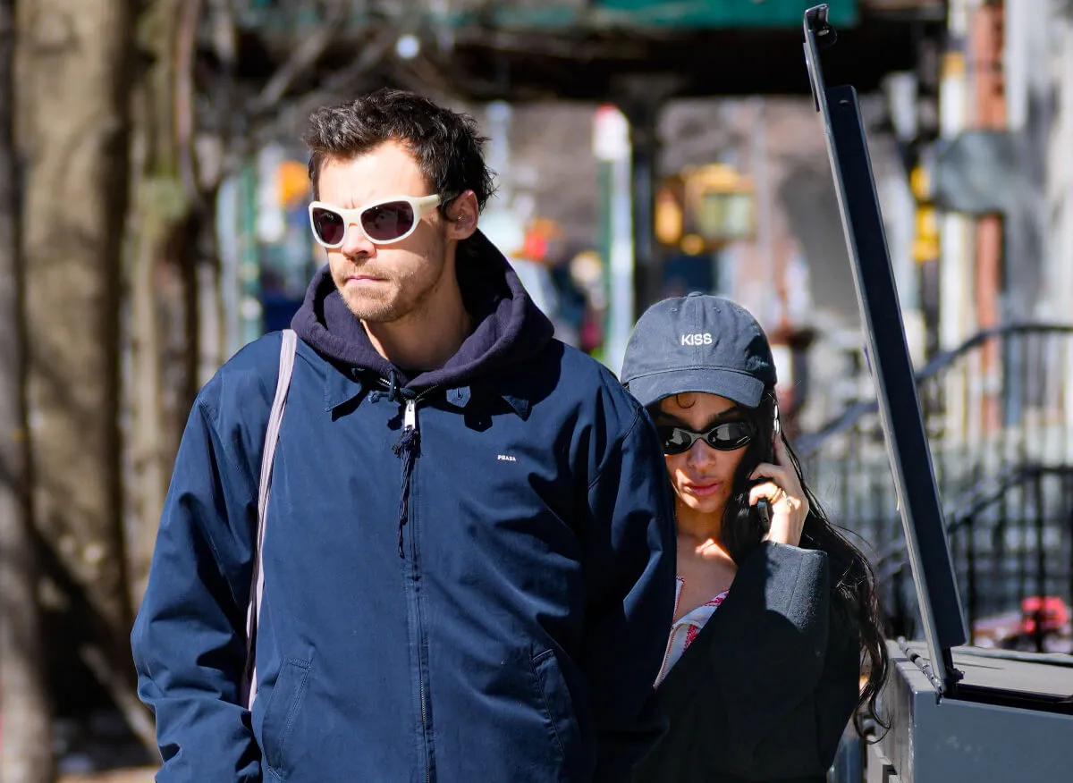 Harry Styles wears a blue coat and sunglasses. He walks with Zoe Kravitz, who wears a hat and sunglasses. She talks on the phone.