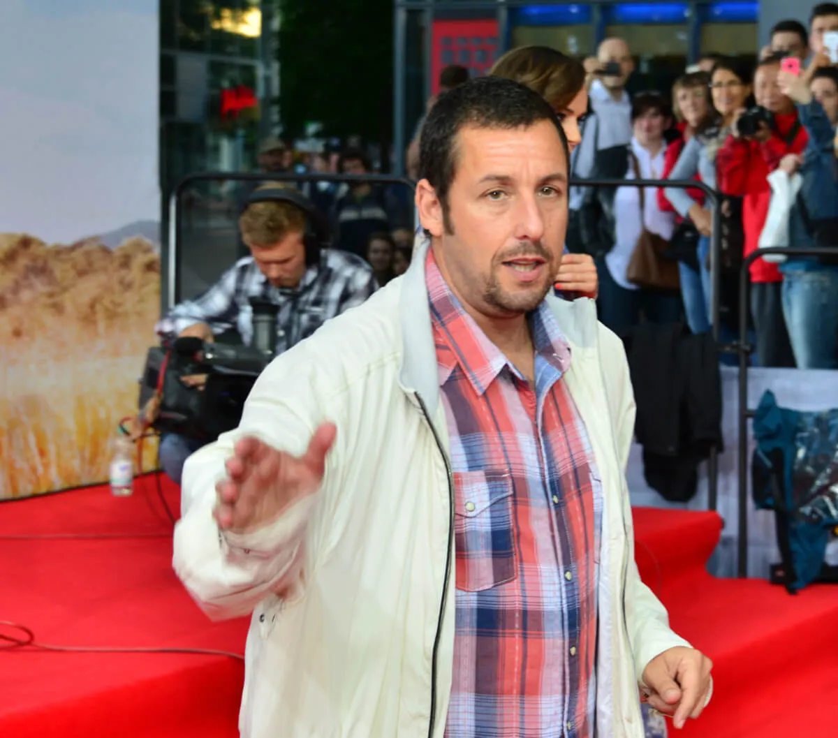 Adam Sandler wears a plaid shirt and a white jacket. He is on the red carpet.