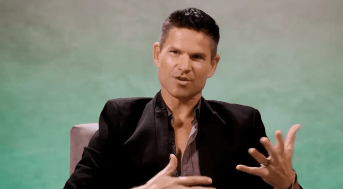 Danny Booko wears a suit jacket and sits in a chair while talking.