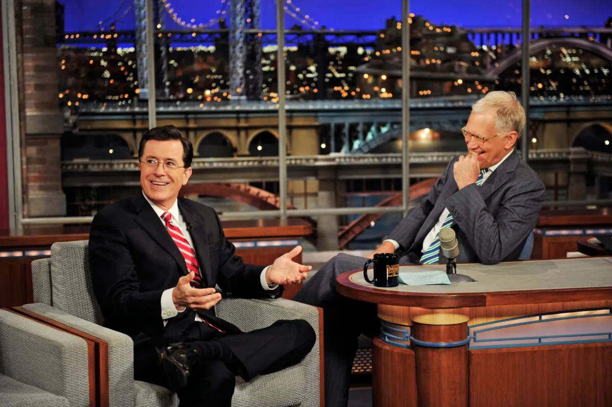 Stephen Colbert sits in an arm chair next to David Letterman's desk.