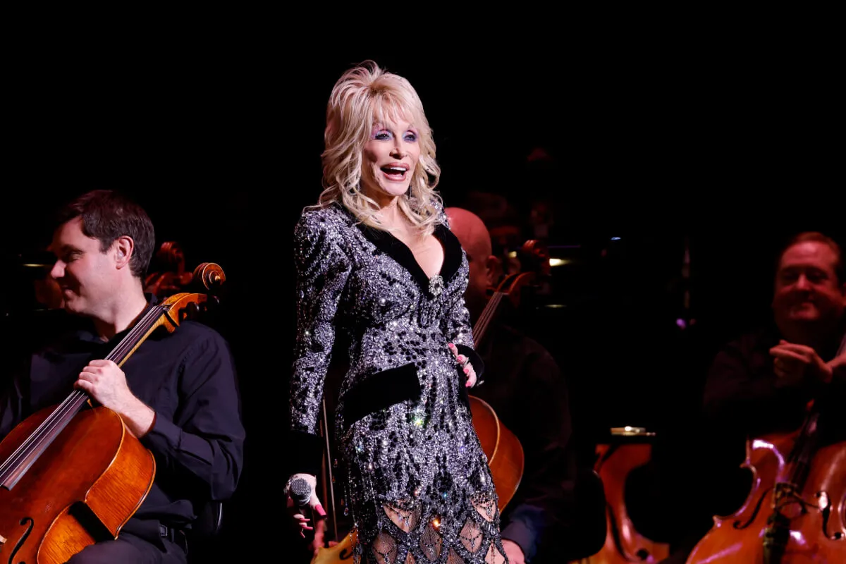 Dolly Parton wears a black dress with silver thread and stands on a stage.
