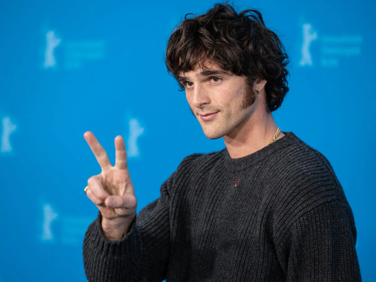Jacob Elordi wears a gray sweater and holds up a peace sign.