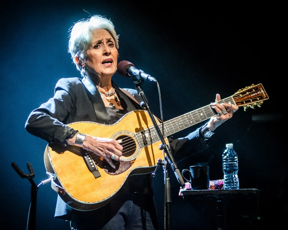 Joan Baez plays a guitar and stands in front of a microphone.