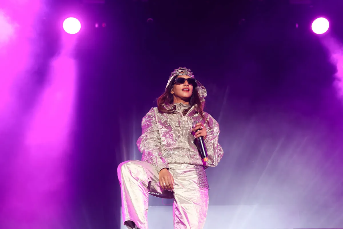 M.I.A. wears silver and sunglasses and stands with a microphone.