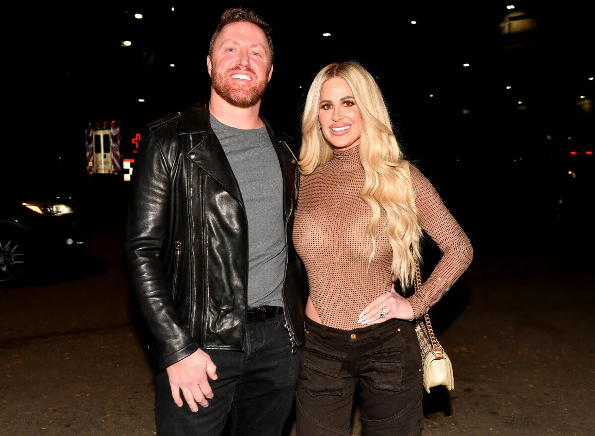 Kim Zolciak wears a brown turtleneck and stands with her hand on her hip. Kroy Biermann wears a black leather jacket.
