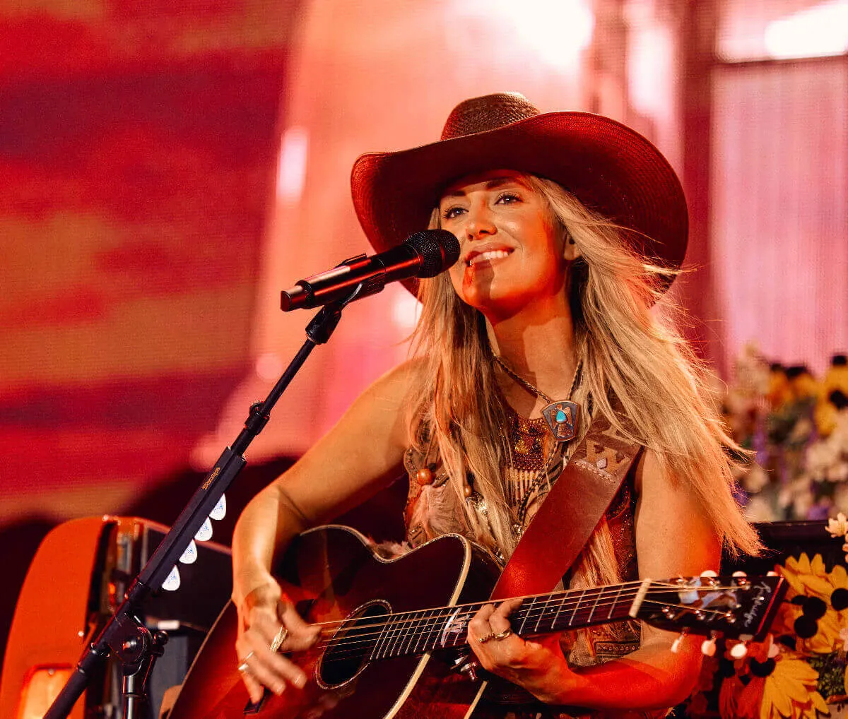 Lainey Wilson wears a cowboy hat and plays guitar in front of a microphone.