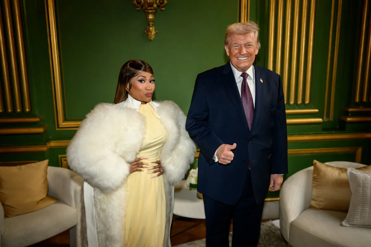 Nicki Minaj wears a yellow dress and a white jacket. She stands next to Donald Trump, who wears a suit.