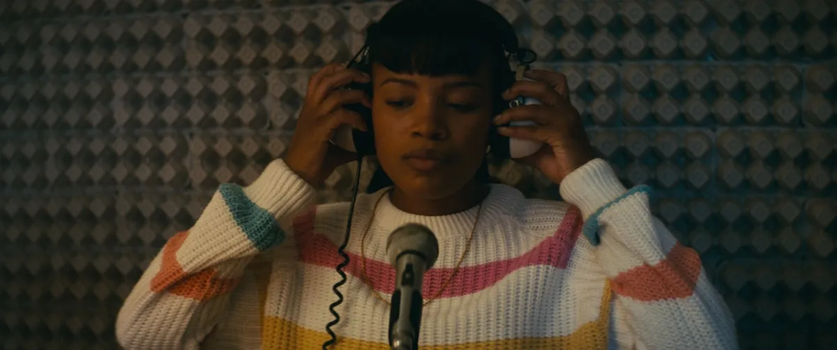 Young woman wearing a striped sweater and headphones singing into a mic in 'Roxanne Roxanne'