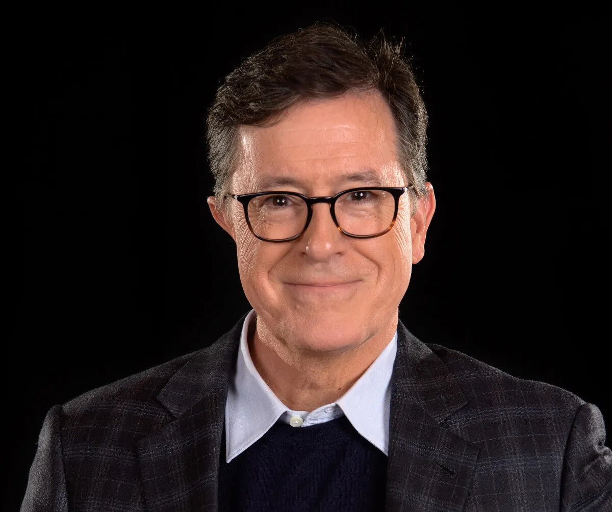 Stephen Colbert wears glasses, a blazer, and a collared shirt.
