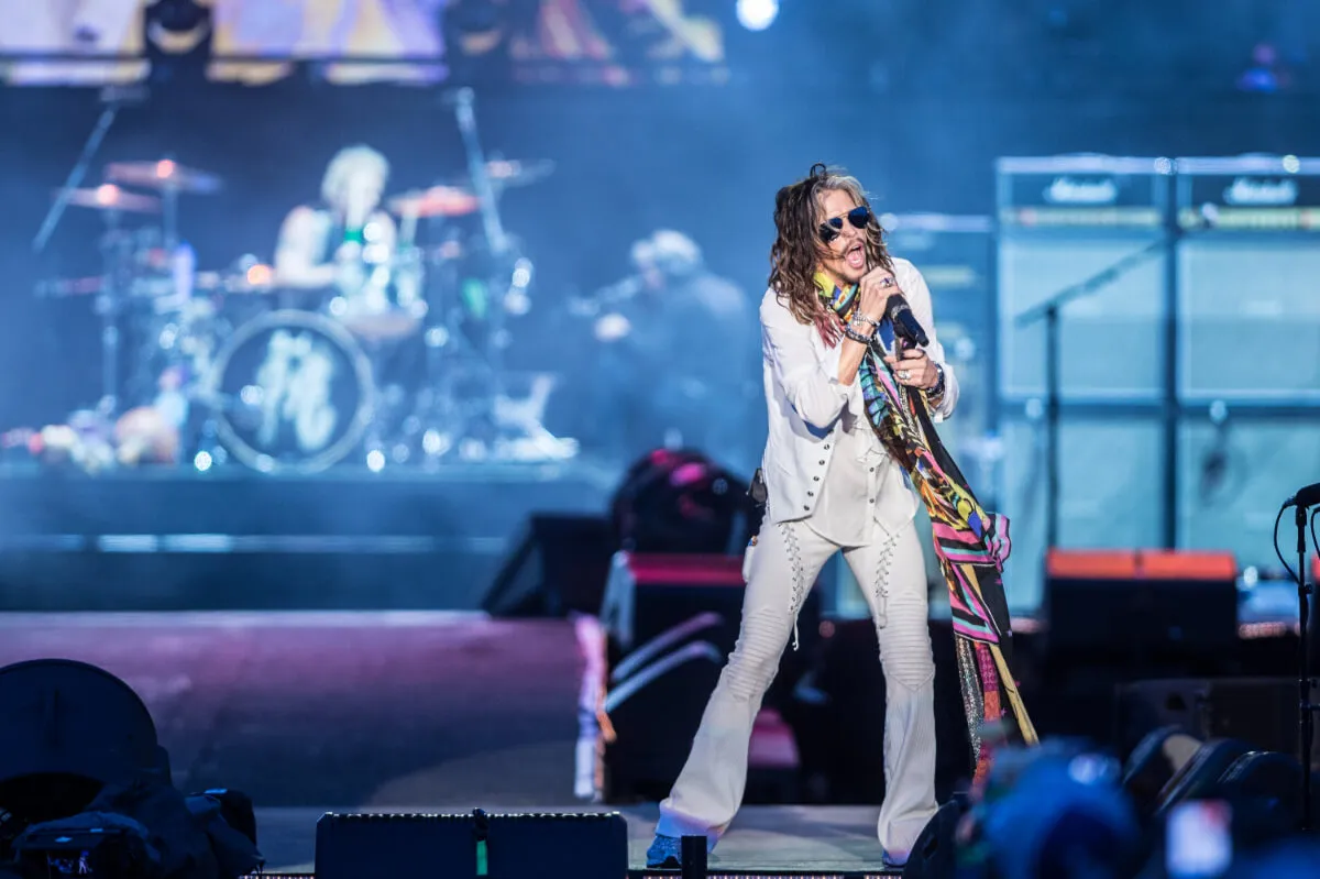 Stephen Tyler wears white and a long, colorful scarf. He sings into a microphone.