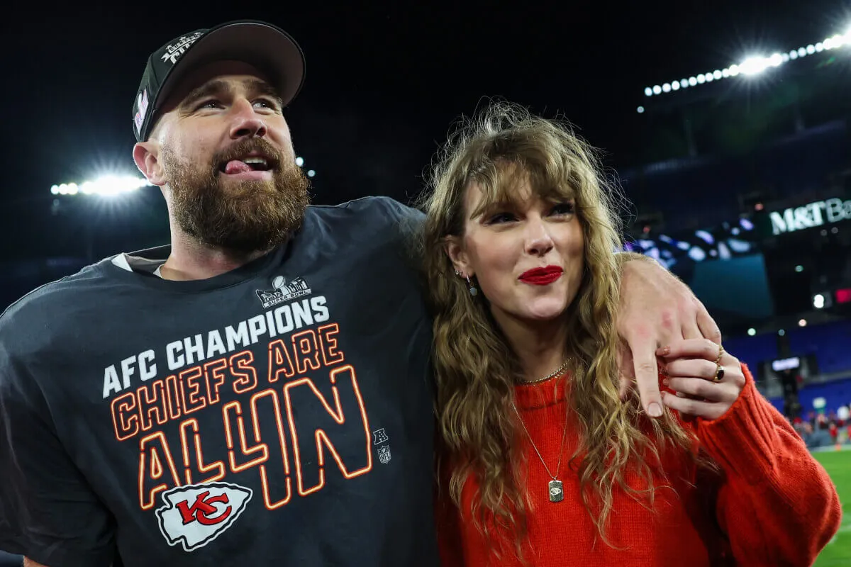 Travis Kelce wears a Chiefs shirt and stands with his arm around Taylor Swift's shoulder. She wears red and holds his hand.