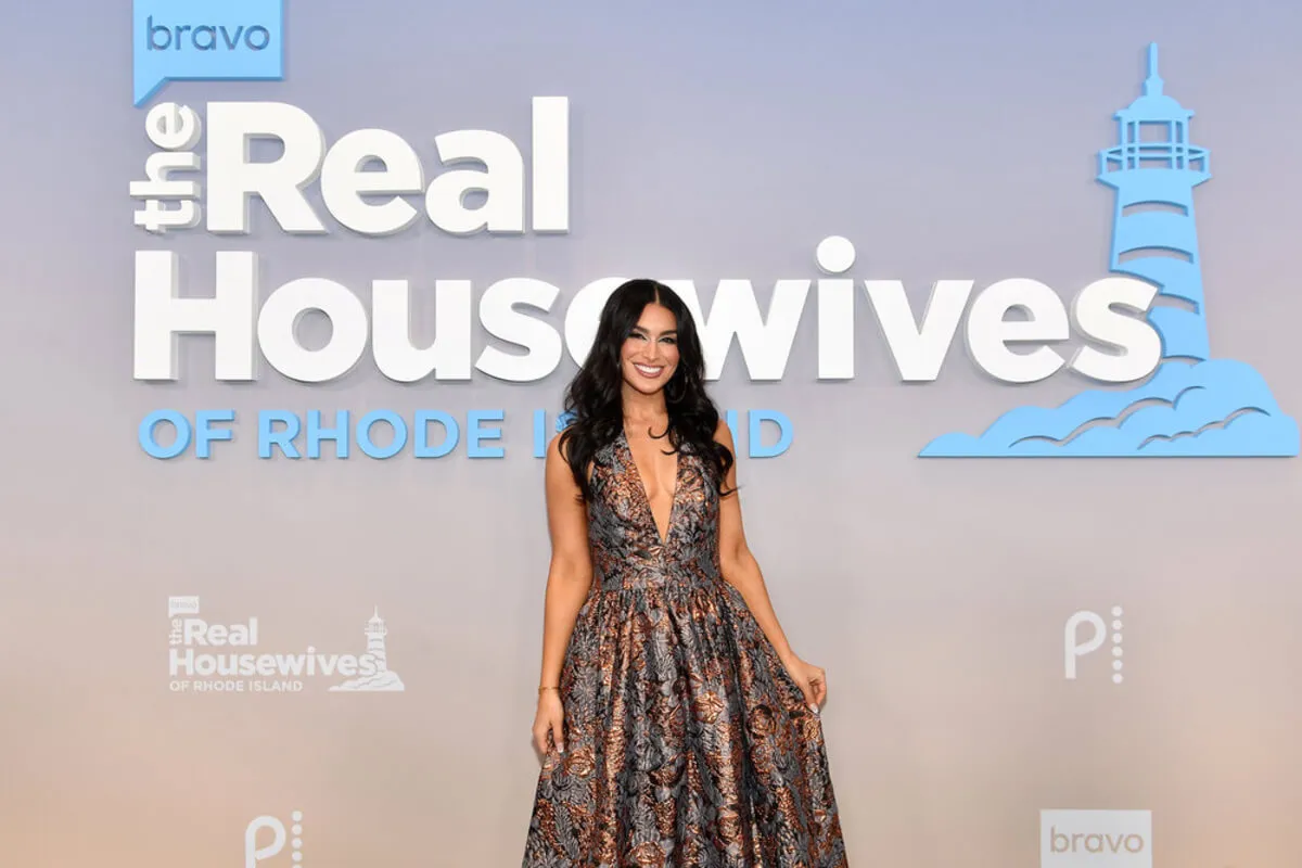 'RHORI' star Ashley Iaconetti wears a dress and stands in front of a sign for the show.