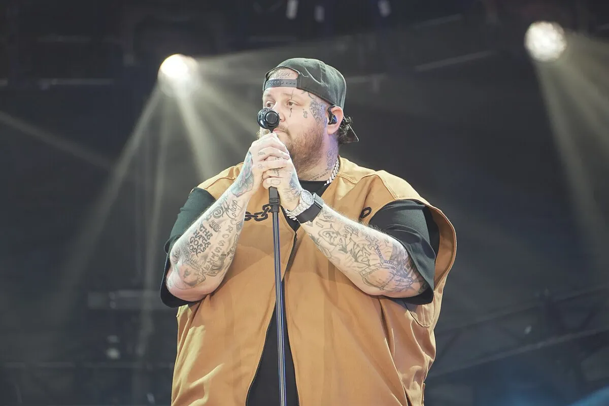 Jelly Roll wears a vest and a backwards baseball hat. He holds a microphone.