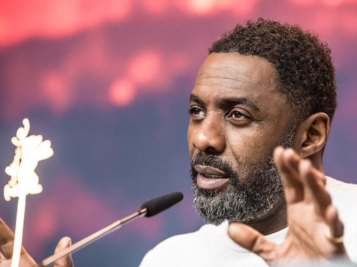 Idris Elba wears a white shirt and speaks into a microphone.