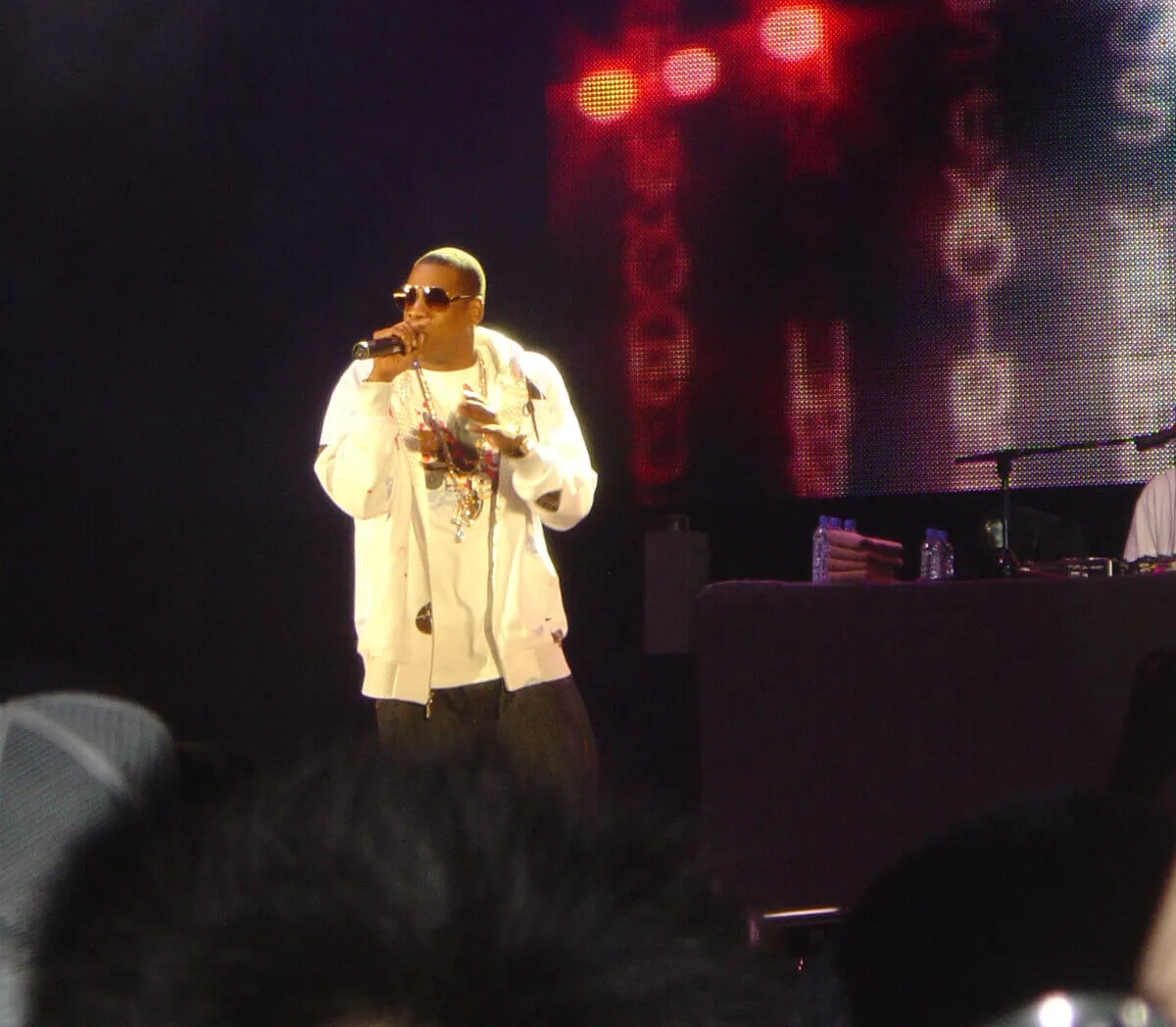 Jay-Z wears white with a pair of sunglasses. He holds a microphone on a stage.