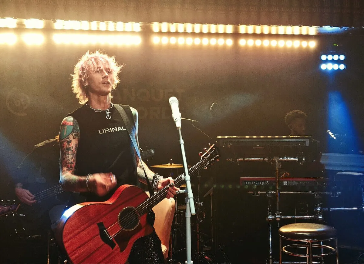 MGK holds an acoustic guitar and stands in front of a microphone.