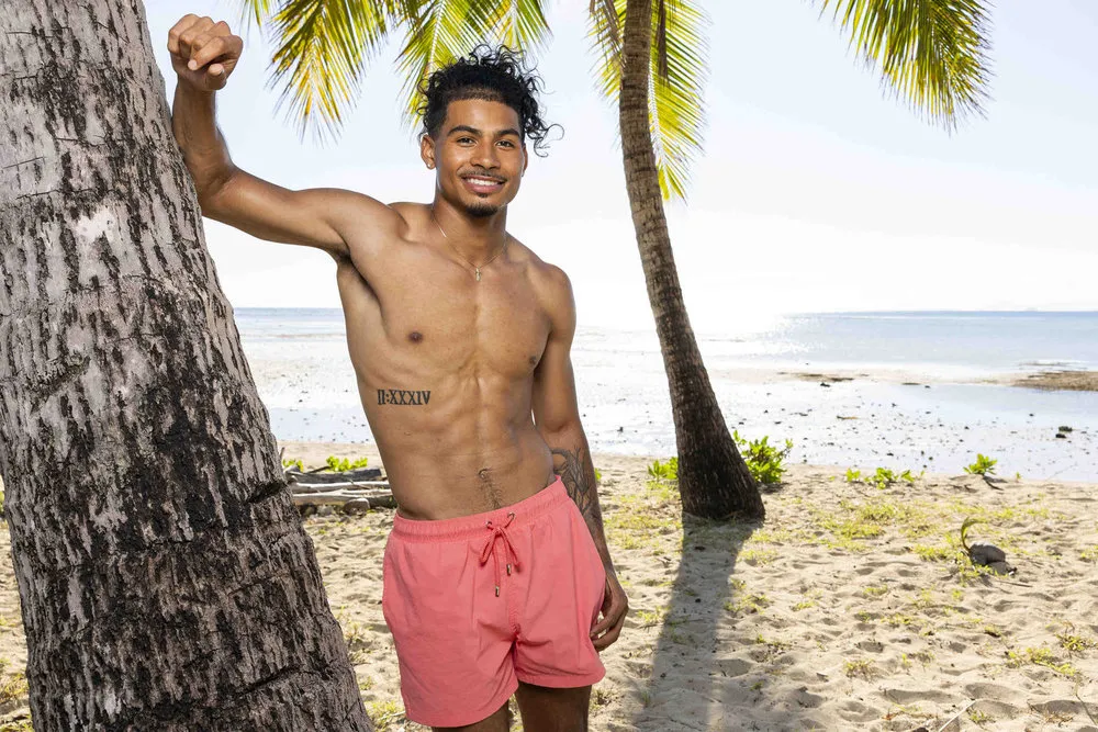 Sincere wears pink swim trunks and leans against a tree on the beach.