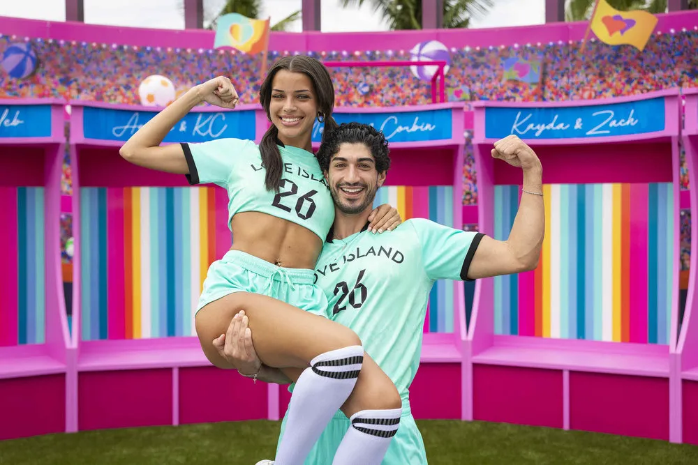 Kayda and Zach wears blue jerseys. He holds her and they both flex a bicep.