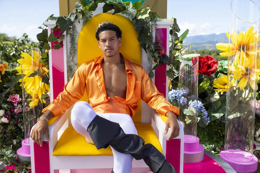 Corbin wears an orange shirt with white pants and sits on a throne.