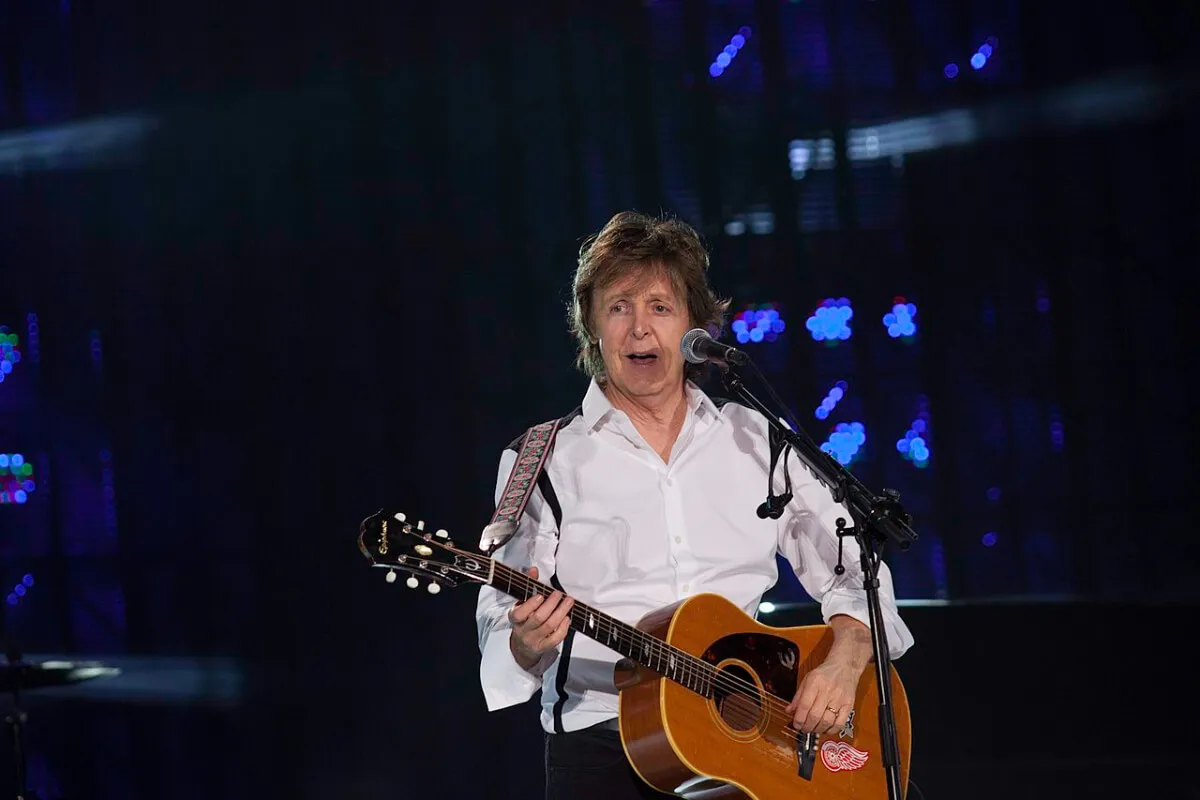 Paul McCartney wears a white shirt and holds an acoustic guitar. He is standing behind a microphone.