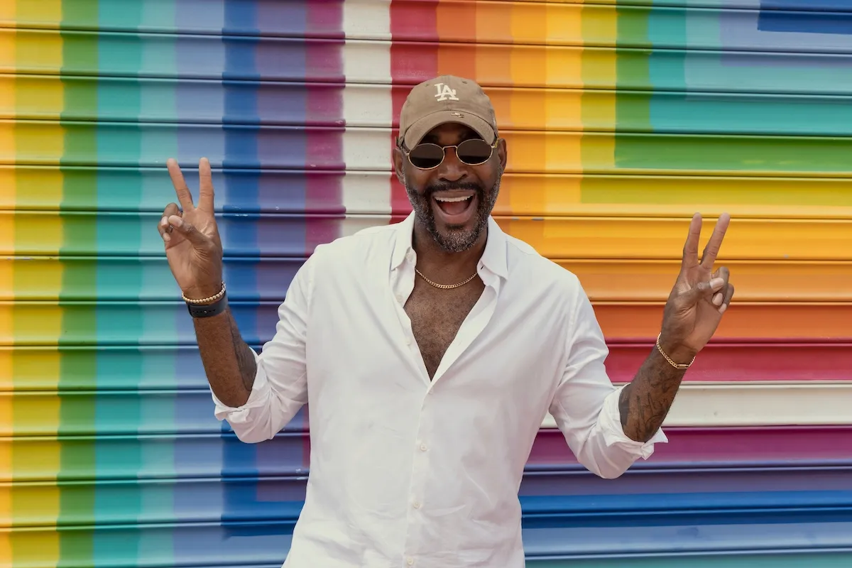 Karamo Brown of 'Queer Eye' wearing a white shirt and standing in front of a rainbow background