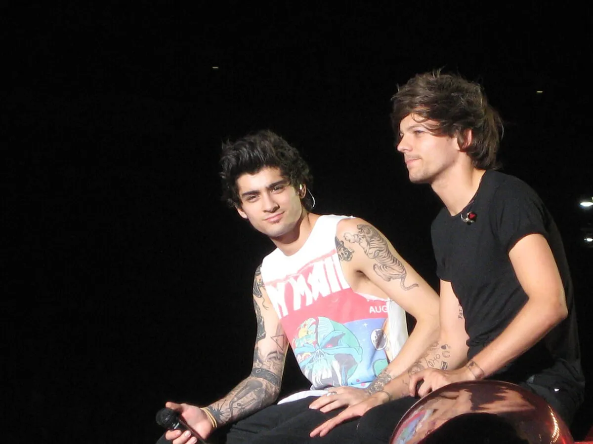 Zayn Malik wears a white tank top and sits next to Louis Tomlinson. He wears a black shirt.