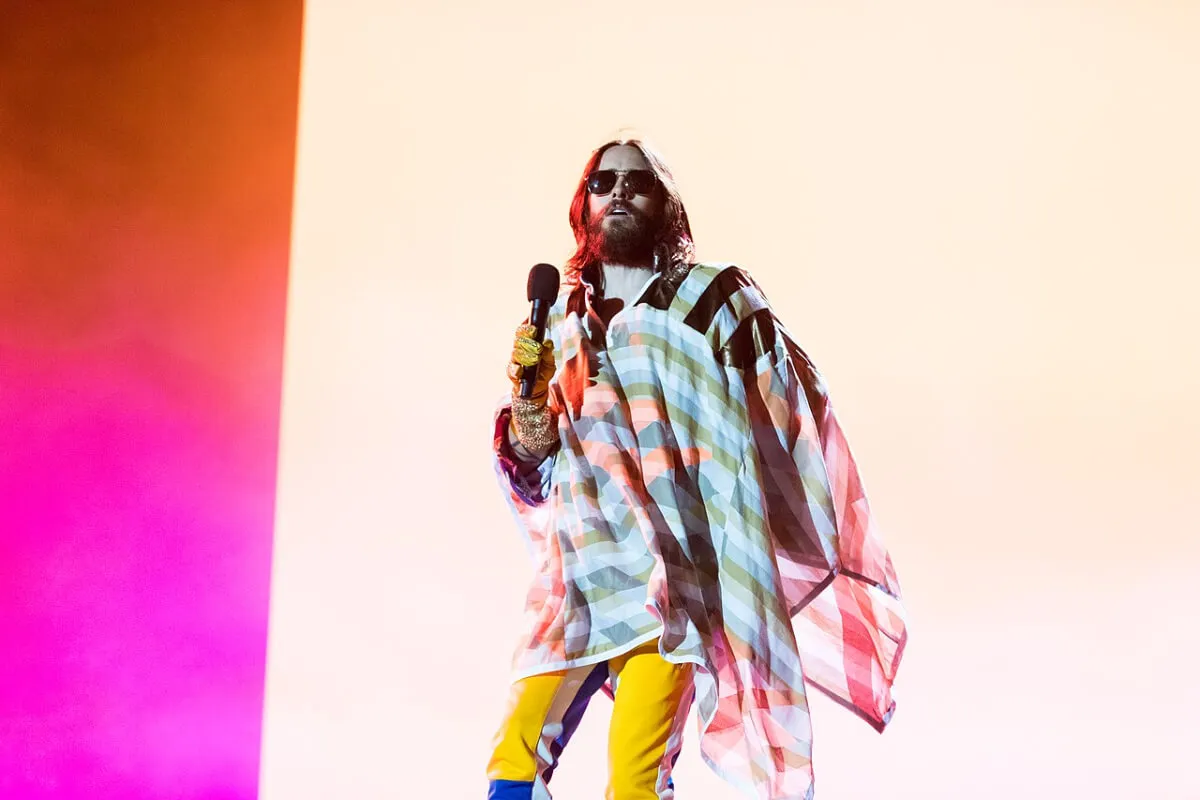 Jared Leto wears sunglasses and a poncho while holding a microphone.