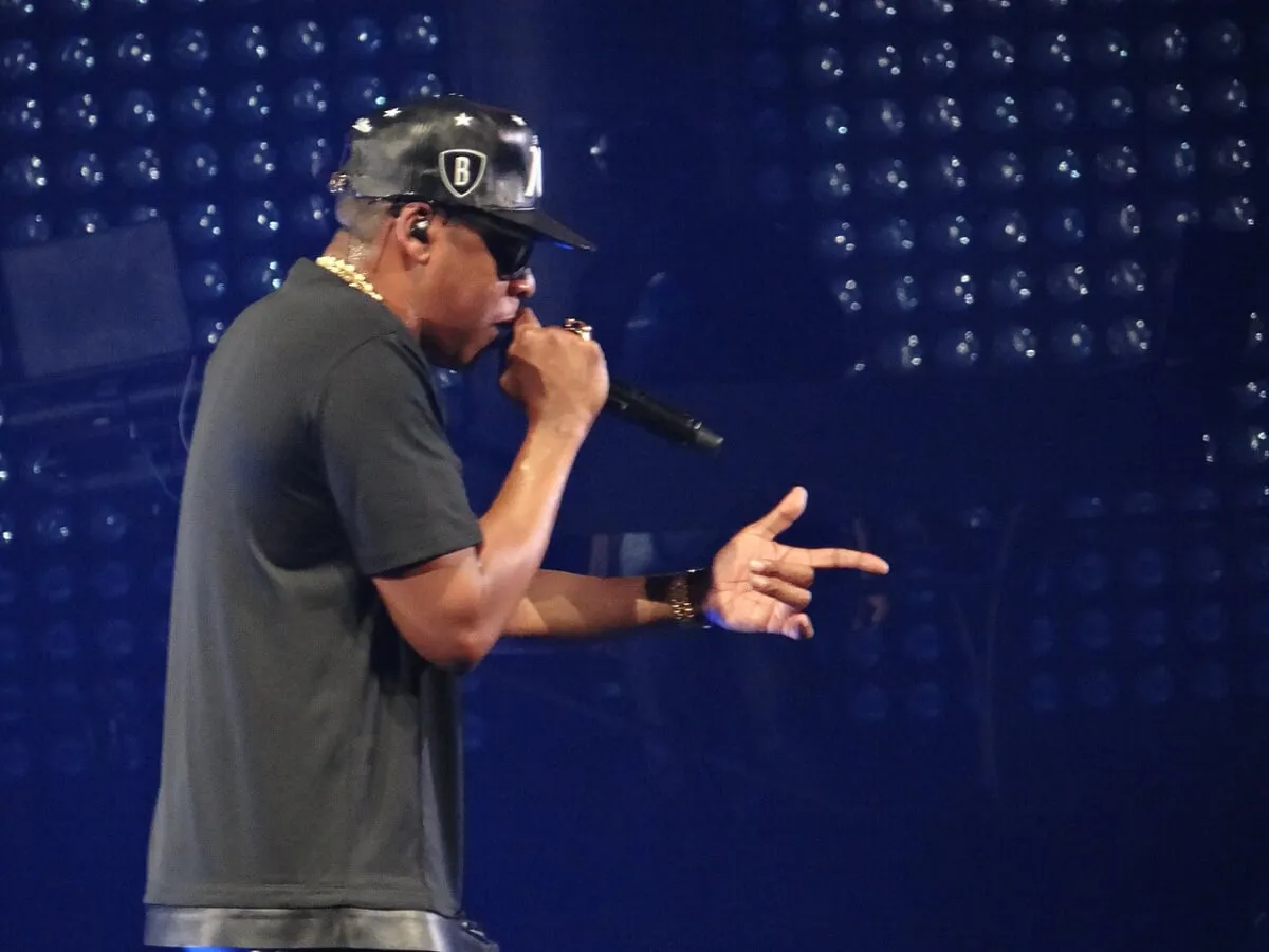 Jay-Z wears a black shirt, hat, and sunglasses. He holds a microphone.