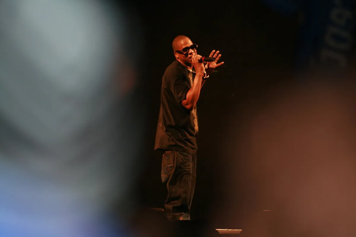 Jay-Z wears a black shirt and sunglasses while on a stage. He holds a microphone.
