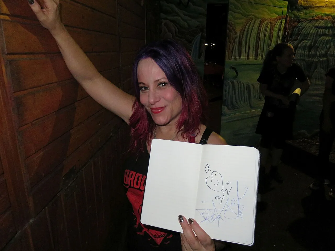 Jennifer Finch, smiling with purple hair, holds an open notebook with a doodle