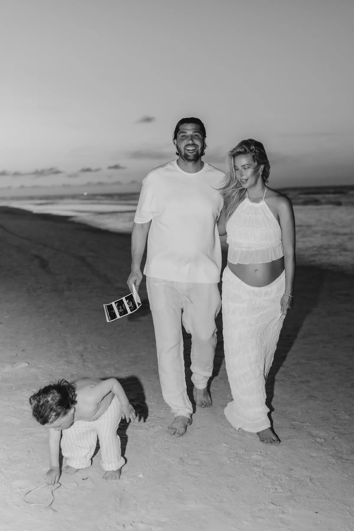 Black and white photo of pregnant Shaina Hurley and her husband walking on a beach with their child in the foreground