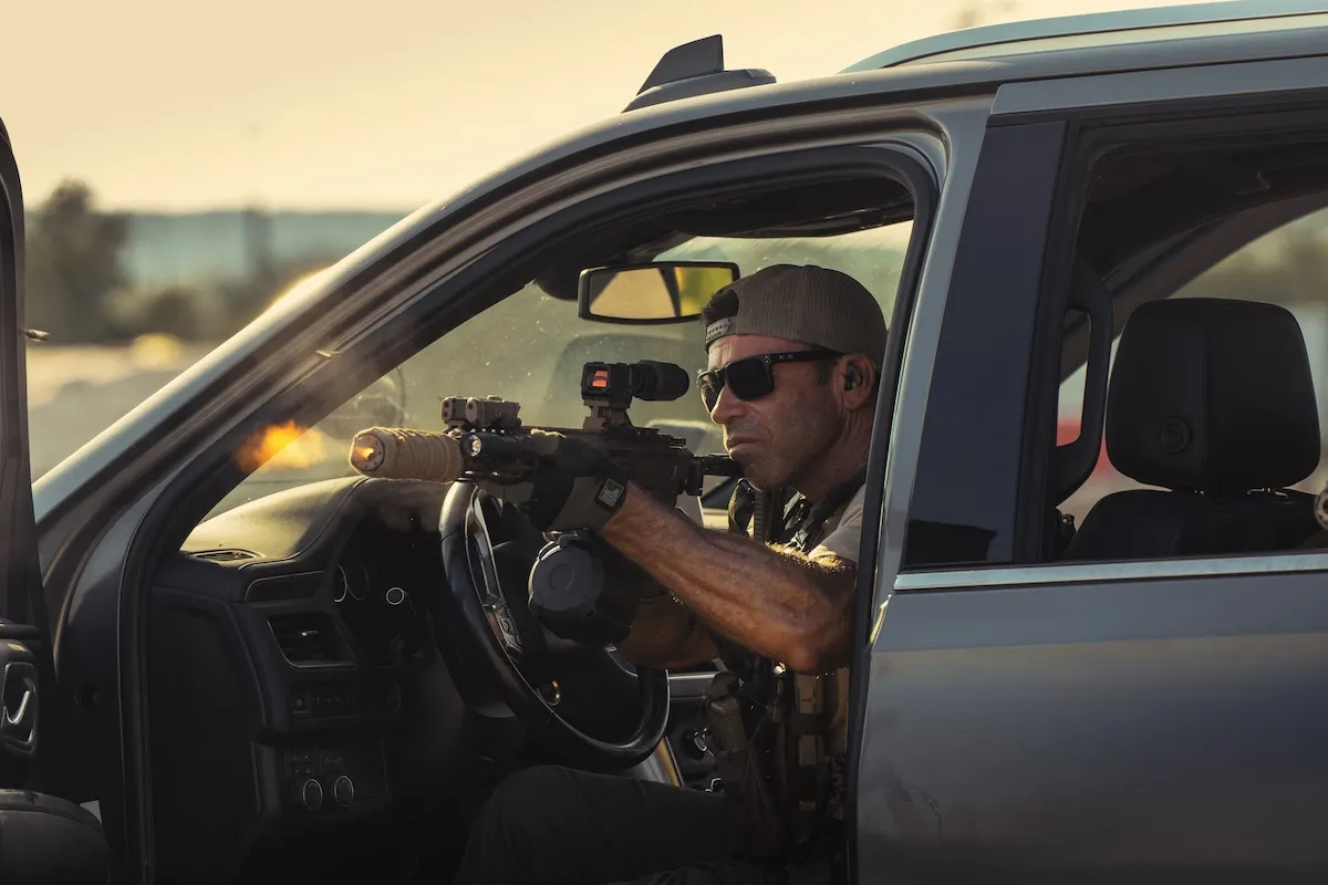 Taylor Sheridan wearing sunglasses and holding a gun while sitting in a car in 'Lioness'