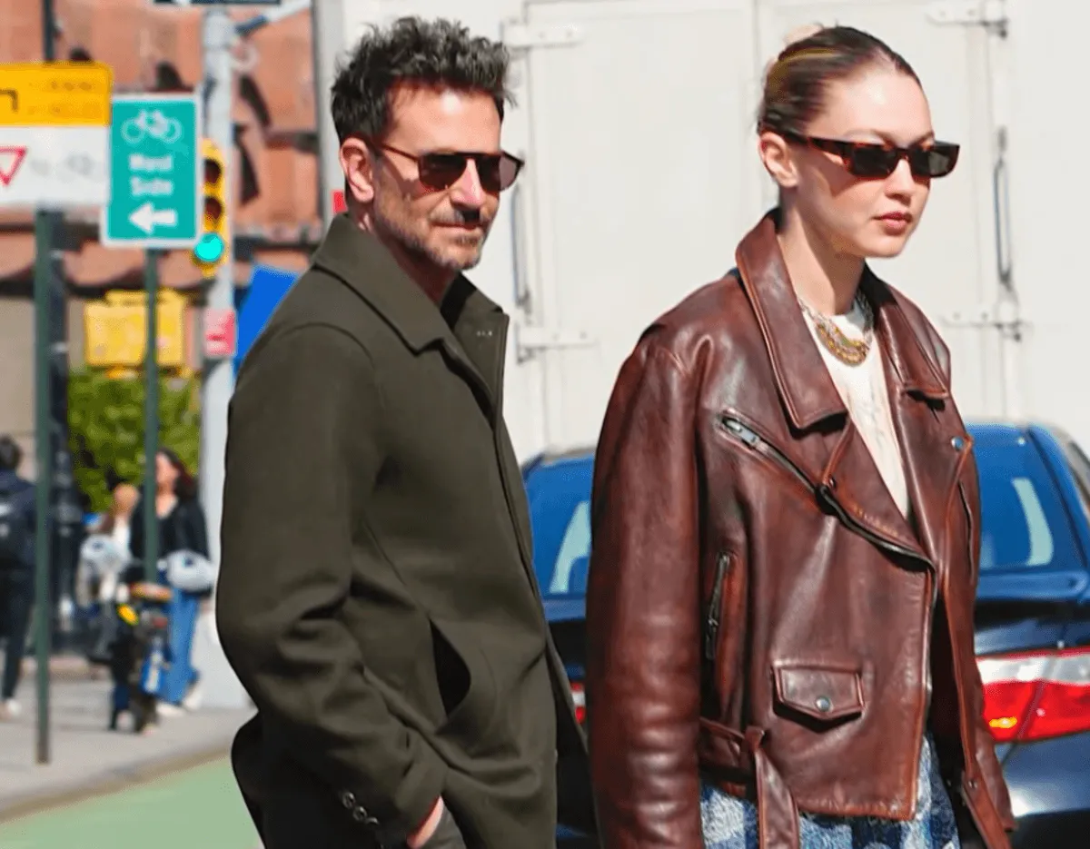 Bradley Cooper wears sunglasses and a green jacket. He walks with Gigi Hadid, who wears sunglasses and a brown leather jacket.