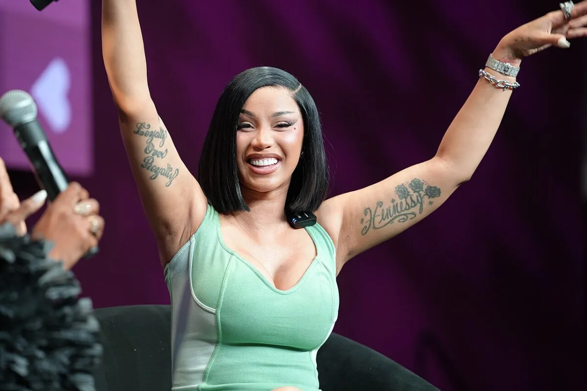 Cardi B wears a green tank top and lifts her arms in the air.