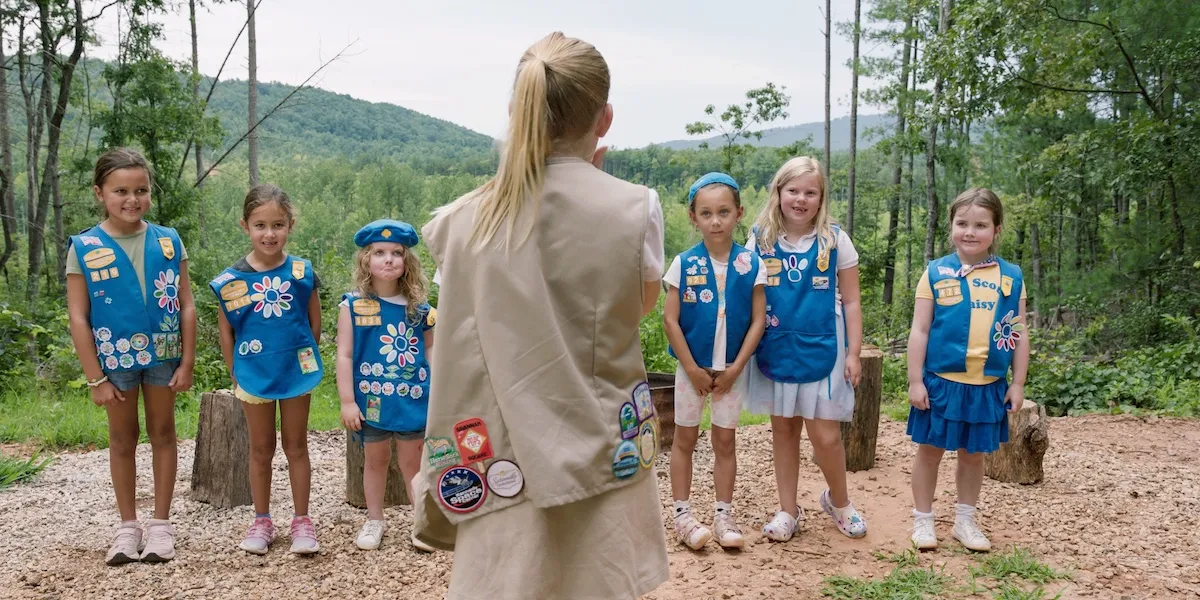 Girl Scout with her back to the camera standing in front of a group of Daisy Girl Scouts in blue vests and smocks