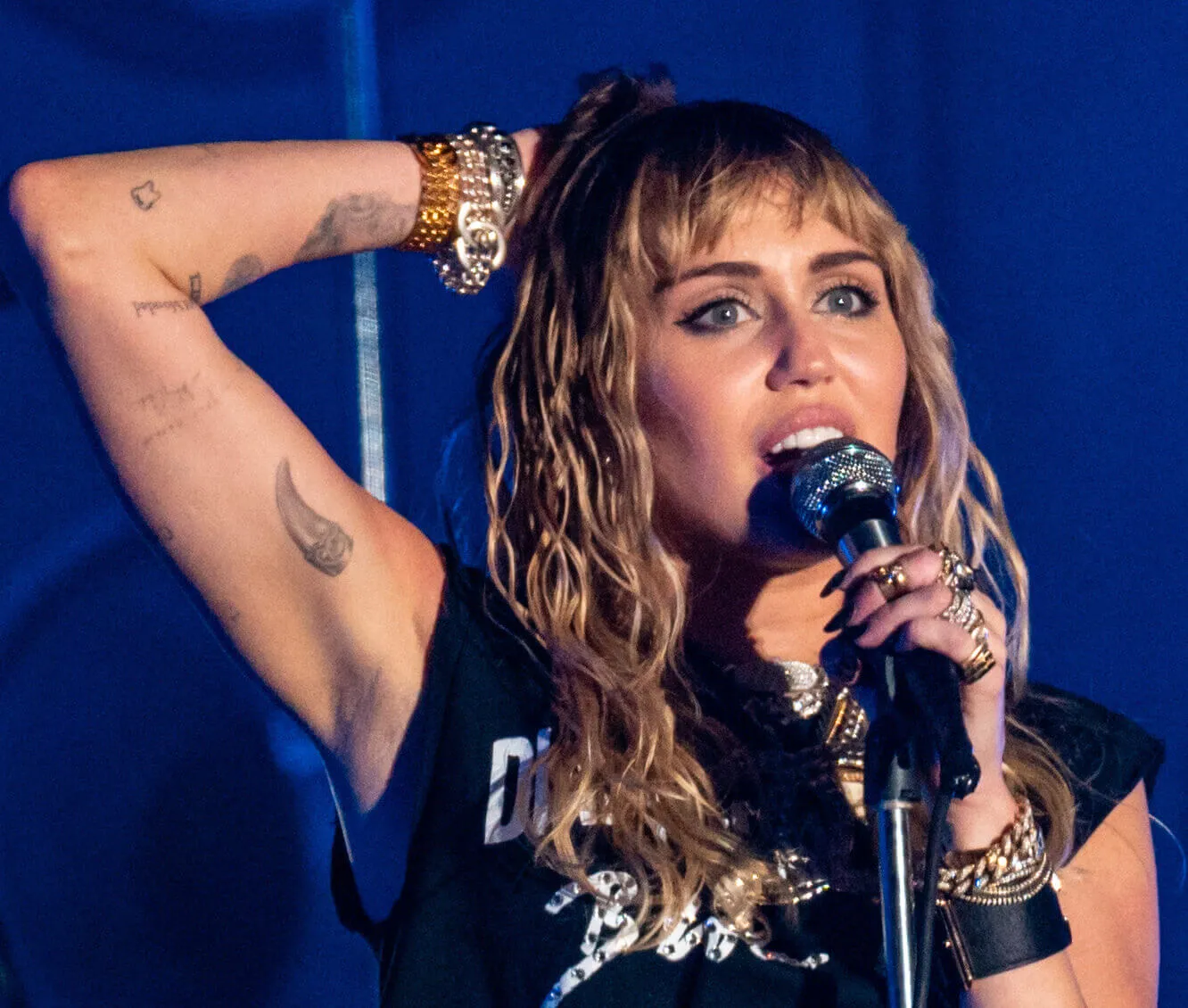 Miley Cyrus wears a black shirt and stands with her hand on her head. She holds a microphone in the other hand.