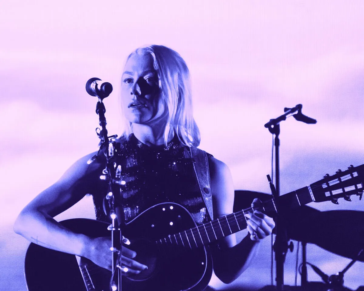 Phoebe Bridgers holds a guitar and stands in front of a microphone.