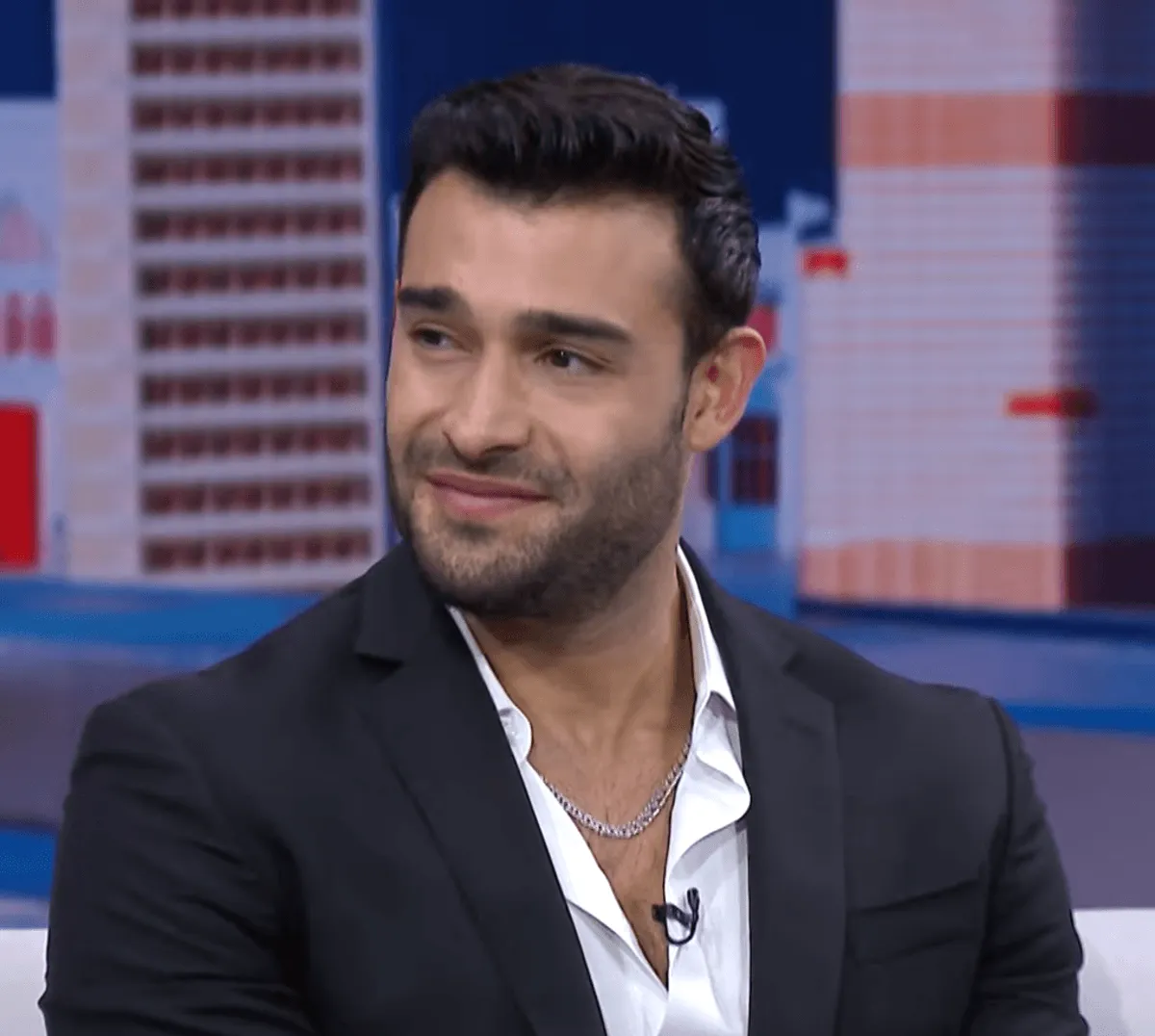 Sam Asghari wears a suit and sits on a couch.