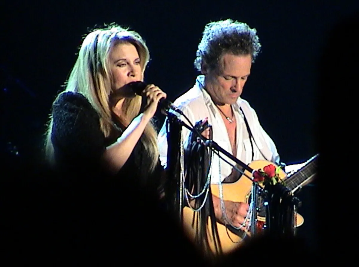 Stevie Nicks and Lindsey Buckingham stand in front of microphones. He wears white and she wears black. He wears guitar.