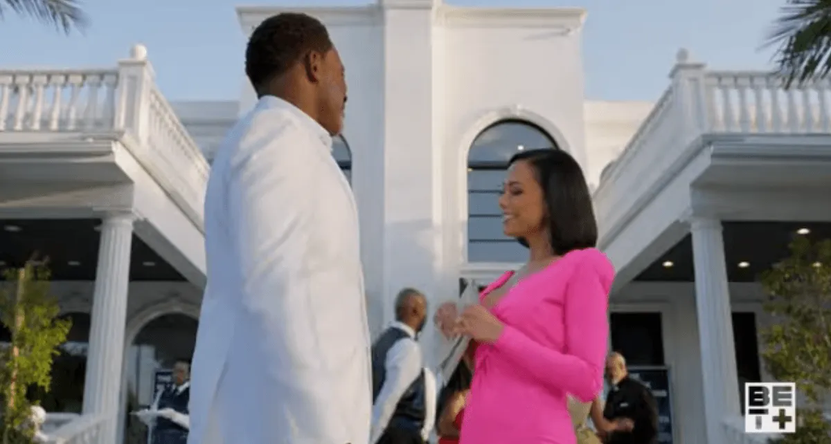 A man dressing in white and a woman in pink looking at each other in front of a white mansion in 'The Black Hamptons'