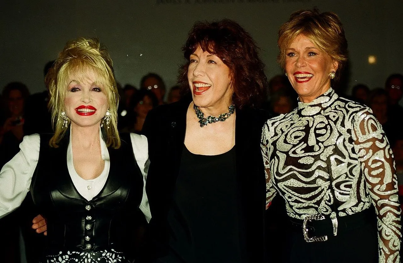 Lily Tomlin stands between Dolly Parton and Jane Fonda. They all smile.