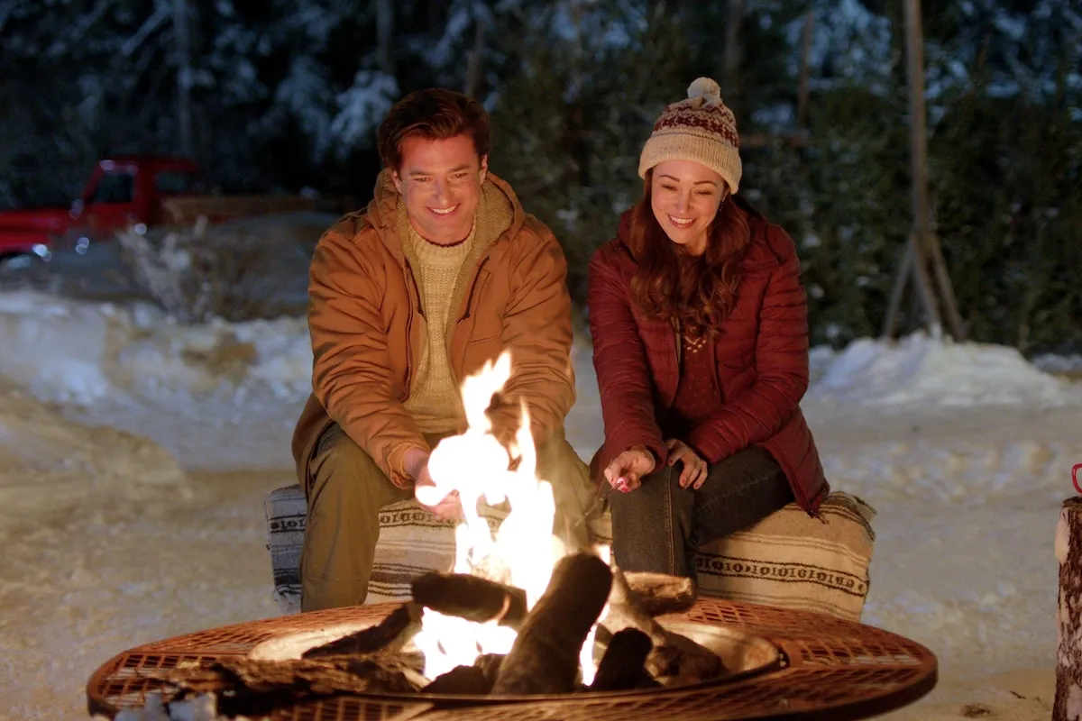 Man and woman dressed in winter coats sitting in front of a camp fire