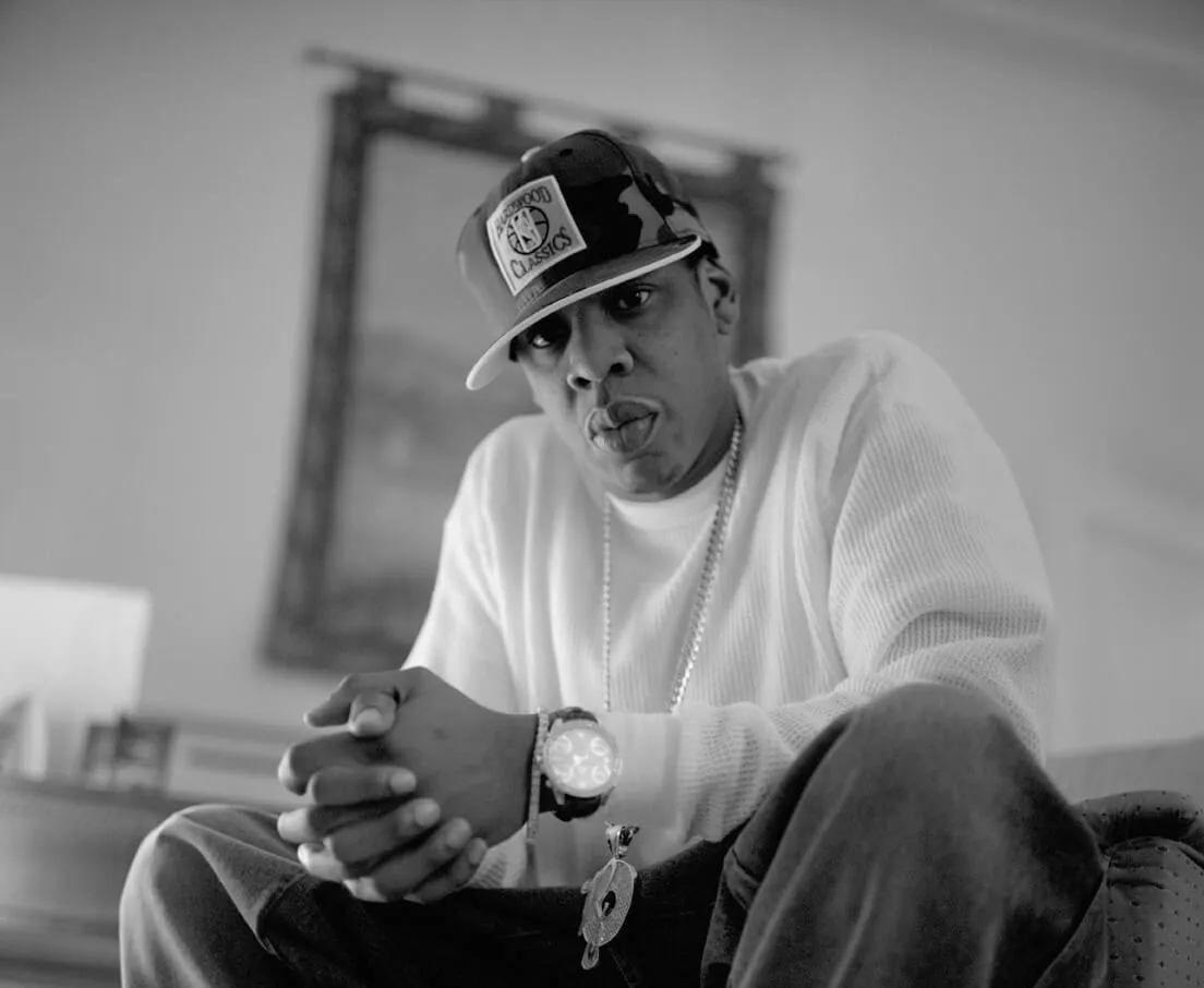 A black and white picture of Jay-Z wearing a hat and sitting with his hands clasped.