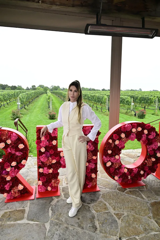 Carole Radziwill wears a cream jumpsuit and stands in front of large red letters. There is a vineyard behind her.