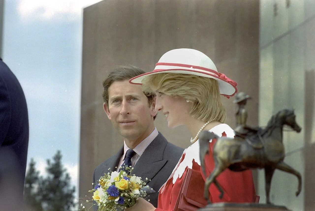 Prince Charles standing next to Princess Diana, wearing a red dress and white hat, and looking at the camera during a royal visit to Canada in 1983