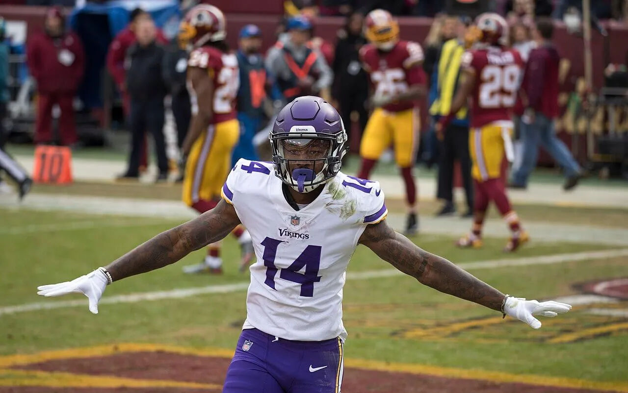 Stefon Diggs wears his football uniform and stands on the field with his arms spread.