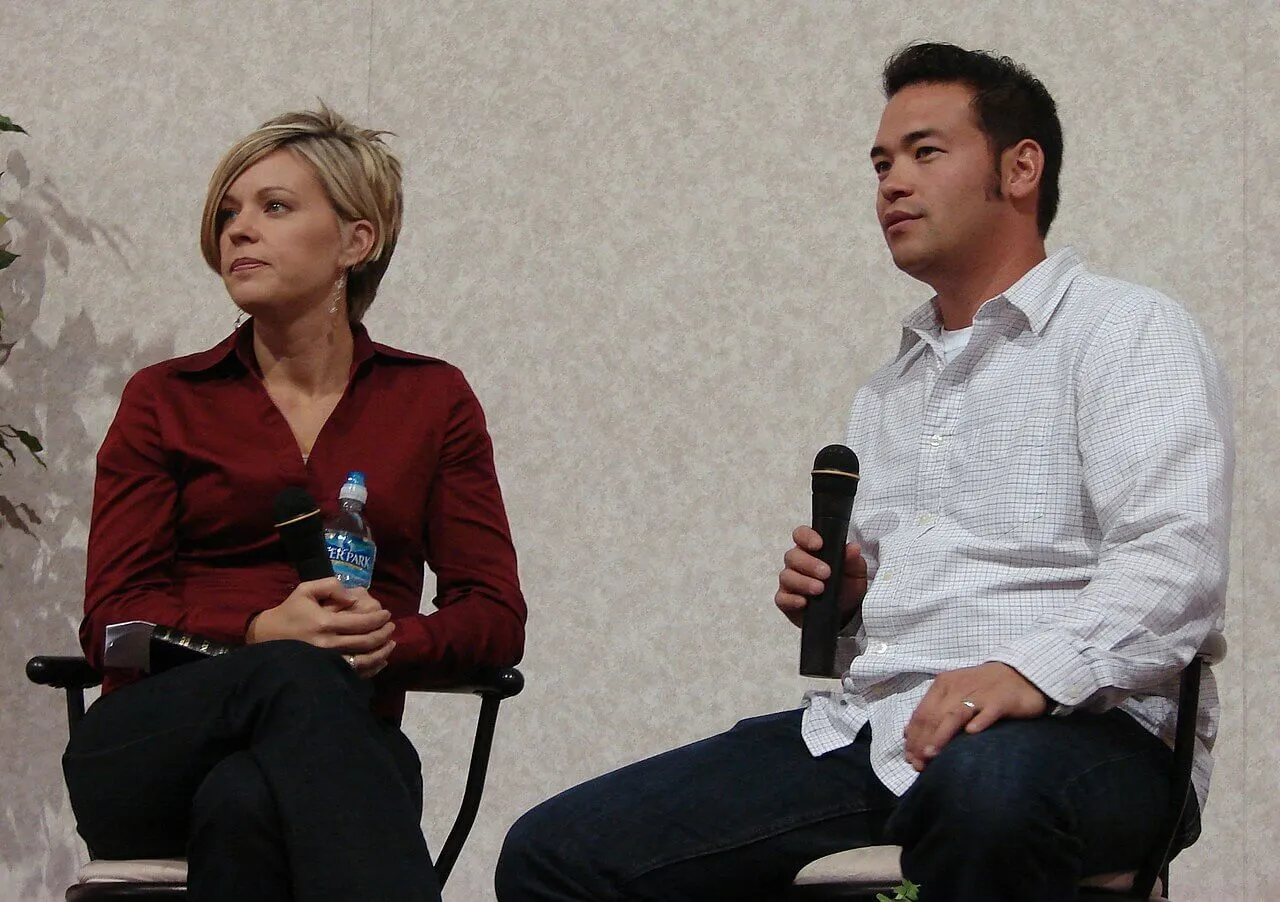 Kate Gosselin wears a red shirt and sits next to Jon Gosselin, who wears a white shirt. She holds a water bottle and he holds a microphone.